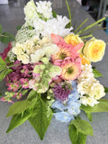 A mixed floral arrangement featuring white, pink, yellow, purple, and light blue flowers with green foliage, photographed on a gray countertop.