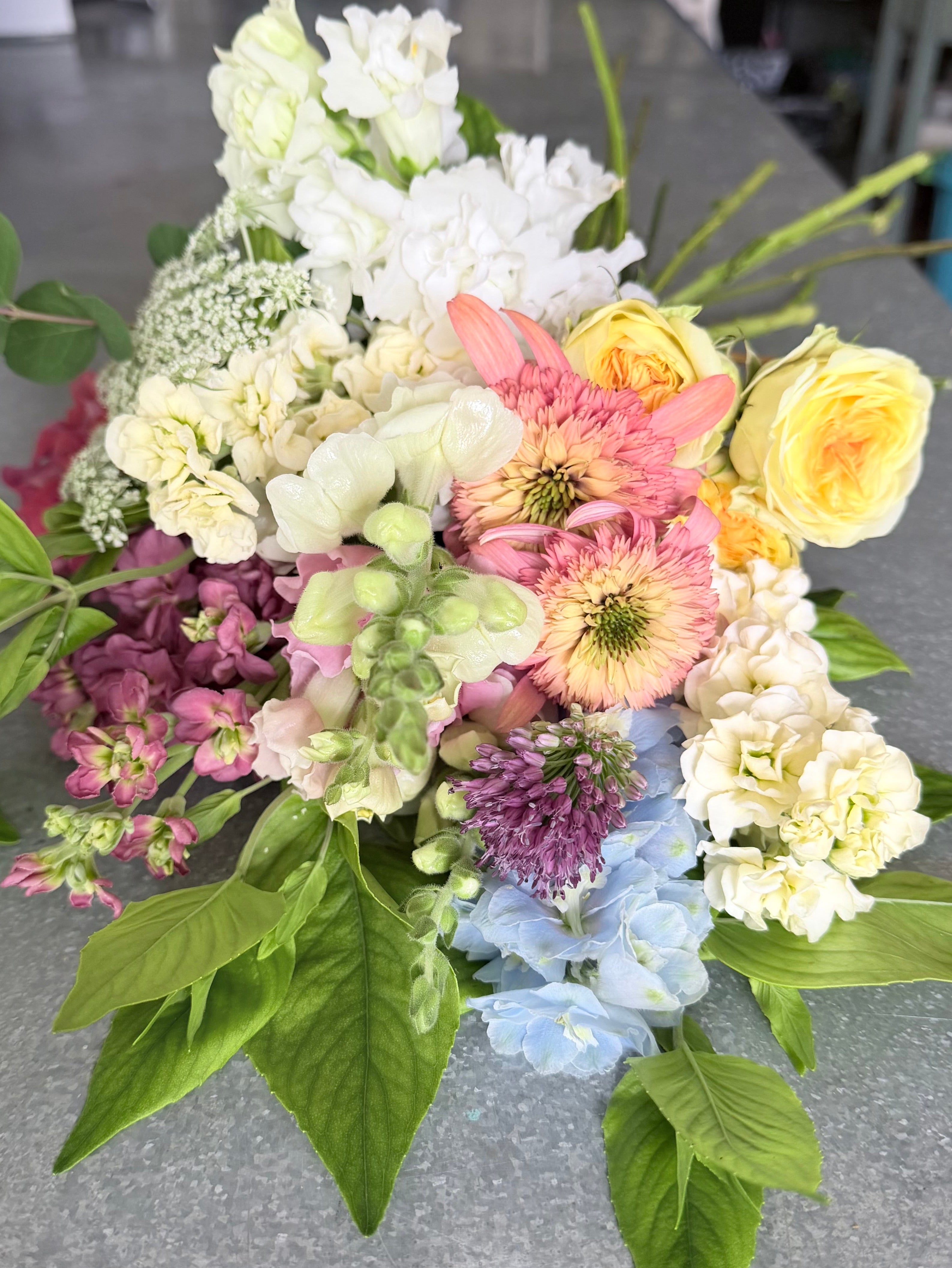 A mixed floral arrangement featuring white, pink, yellow, purple, and light blue flowers with green foliage, photographed on a gray countertop.
