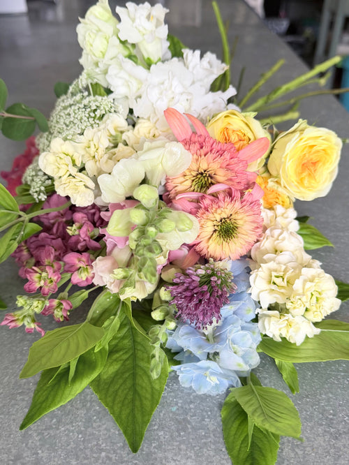 A mixed floral arrangement featuring white, pink, yellow, purple, and light blue flowers with green foliage, photographed on a gray countertop.